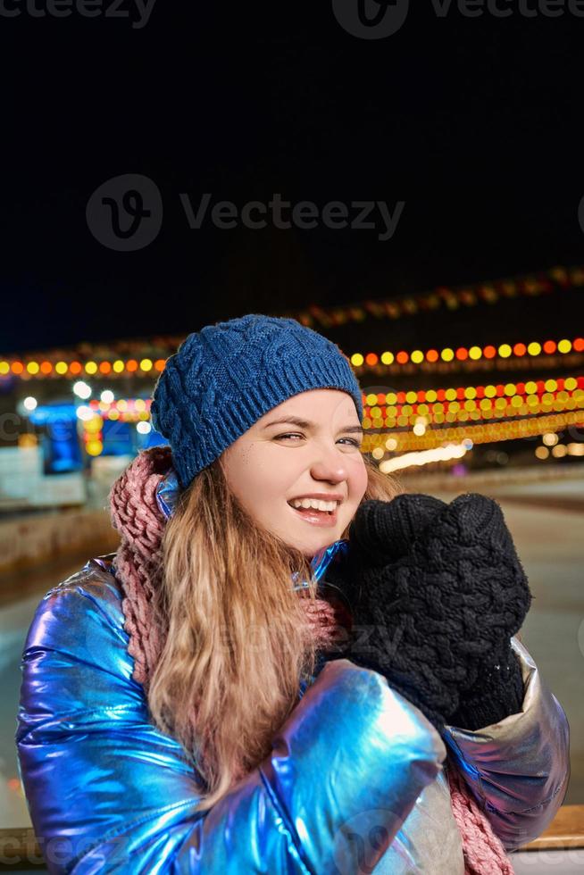 felice sorridente giovane donna caucasica in sciarpa, cappello, giacca, guanti sulla pista di pattinaggio all'aperto. anno nuovo, divertimento, concetto invernale foto