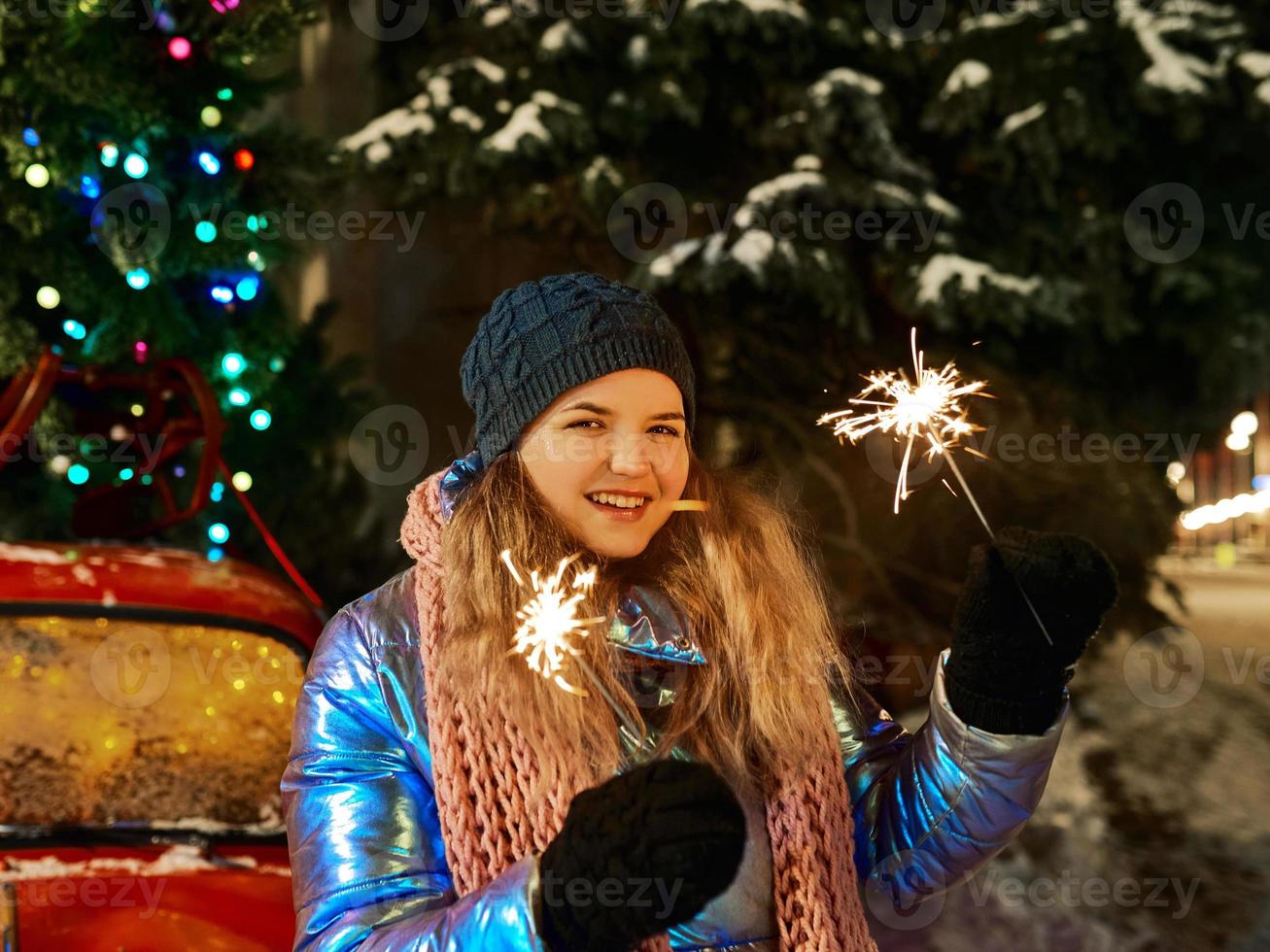 felice sorridente giovane donna caucasica in sciarpa, cappello, giacca, guanti con sparkler dall'albero di Natale all'aperto. anno nuovo, divertimento, concetto invernale foto