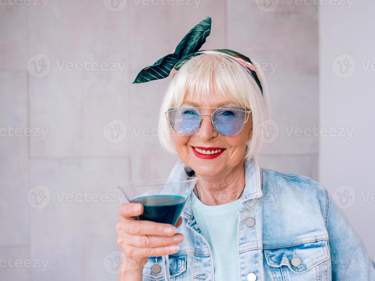 donna anziana alla moda con i capelli grigi e con occhiali blu e giacca di jeans che tiene in mano un bicchiere con cocktail blu. alcol, relax, vacanze, concetto di pensione foto