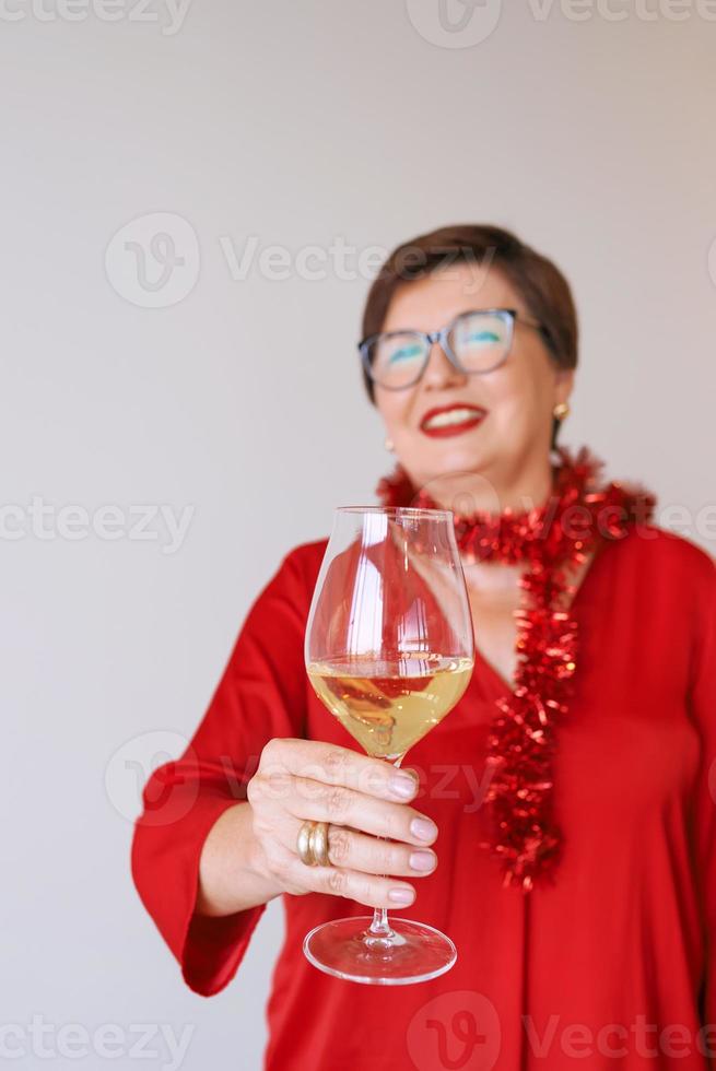 elegante donna anziana matura in camicetta rossa con un bicchiere di vino bianco per celebrare il nuovo anno. divertimento, festa, stile, stile di vita, alcol, concetto di celebrazione foto