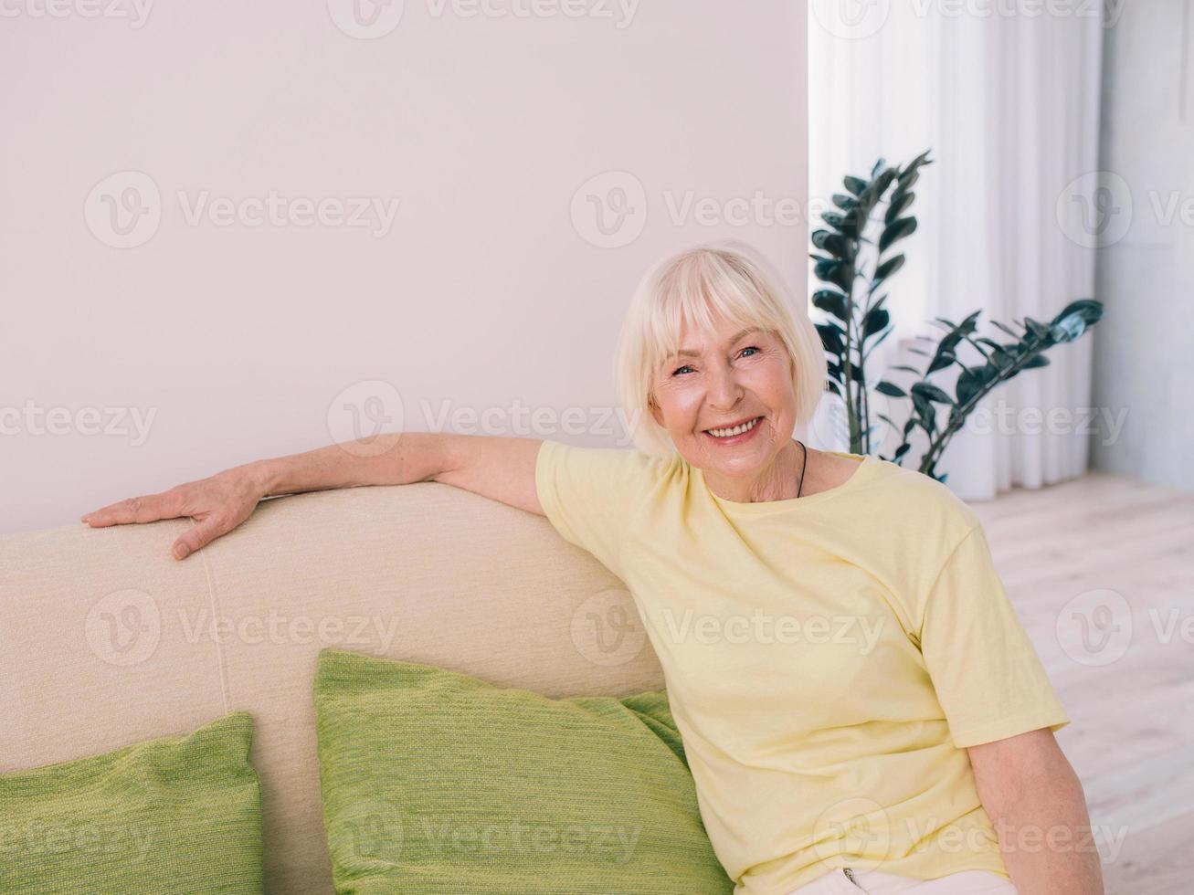 allegra donna caucasica elegante con i capelli grigi seduto sul divano di casa. anti età, stile di vita sano, concetto di pensiero positivo foto