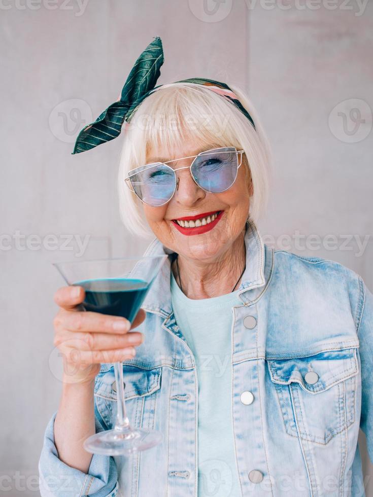 donna anziana alla moda con i capelli grigi e con occhiali blu e giacca di jeans che tiene in mano un bicchiere con cocktail blu. alcol, relax, vacanze, concetto di pensione foto