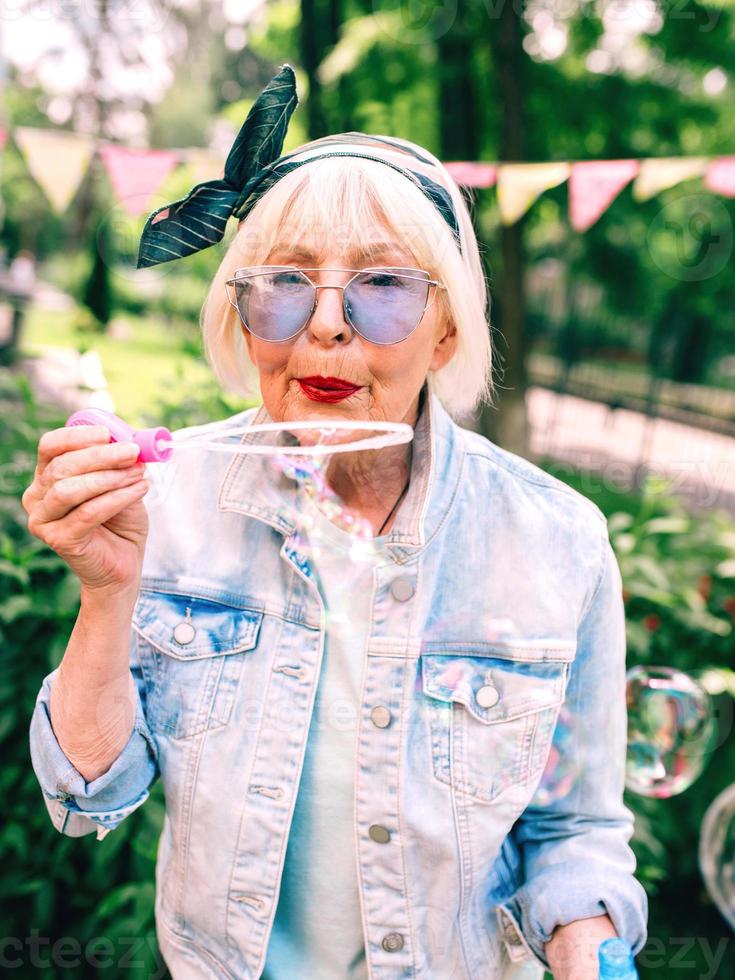 donna alla moda senior con capelli grigi e in occhiali blu e giacca di jeans che soffia bolle all'aperto. vacanze, festa, anti età, concetto di divertimento foto