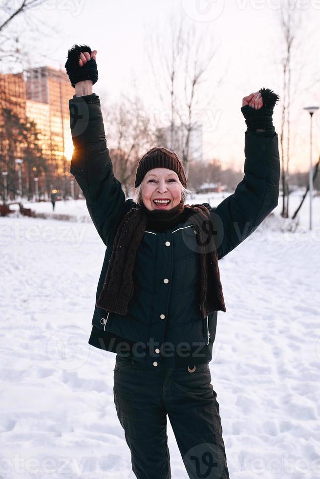 donna anziana in cappello e giacca sportiva che fa esercizi sportivi nel parco invernale di neve. inverno, età, sport, attività, concetto di stagione foto