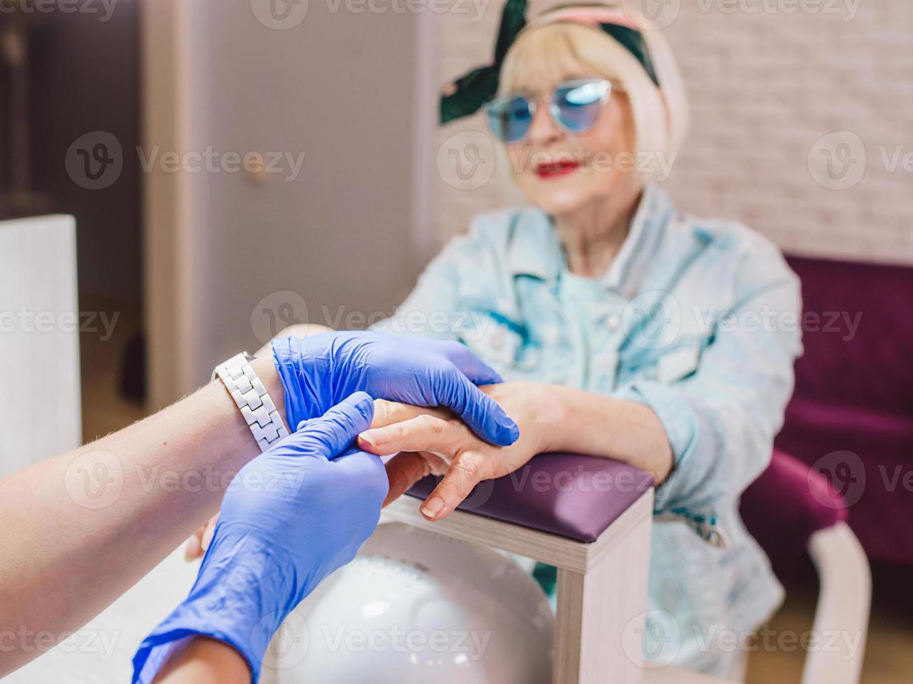 maestro di manicure in guanti blu che screma le mani di una donna anziana alla moda con occhiali da sole blu e giacca di jeans seduta al salone di manicure foto