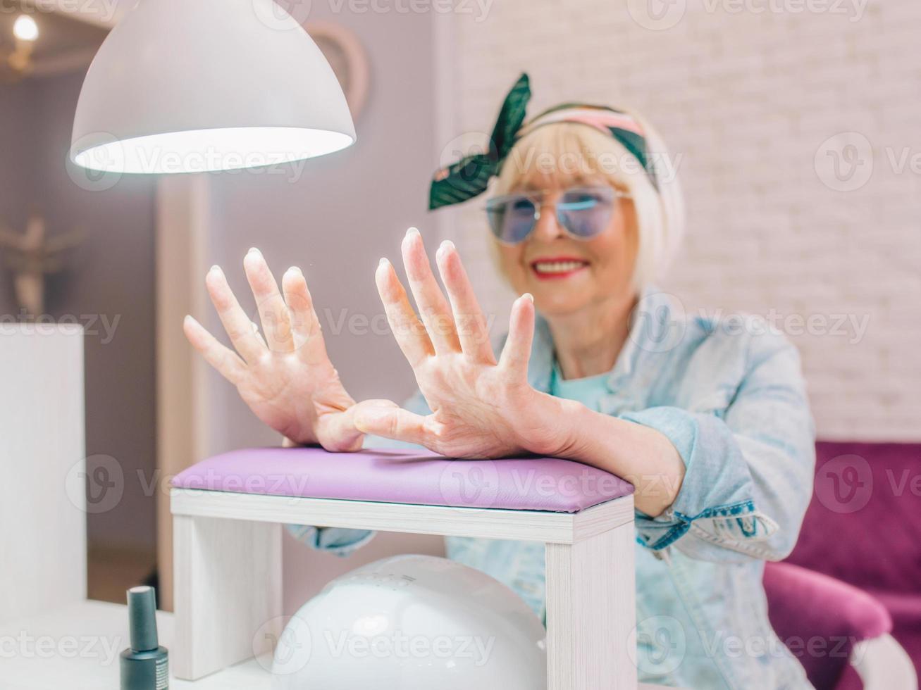 maestro di manicure in guanti blu che screma le mani di una donna anziana alla moda con occhiali da sole blu e giacca di jeans seduta al salone di manicure foto