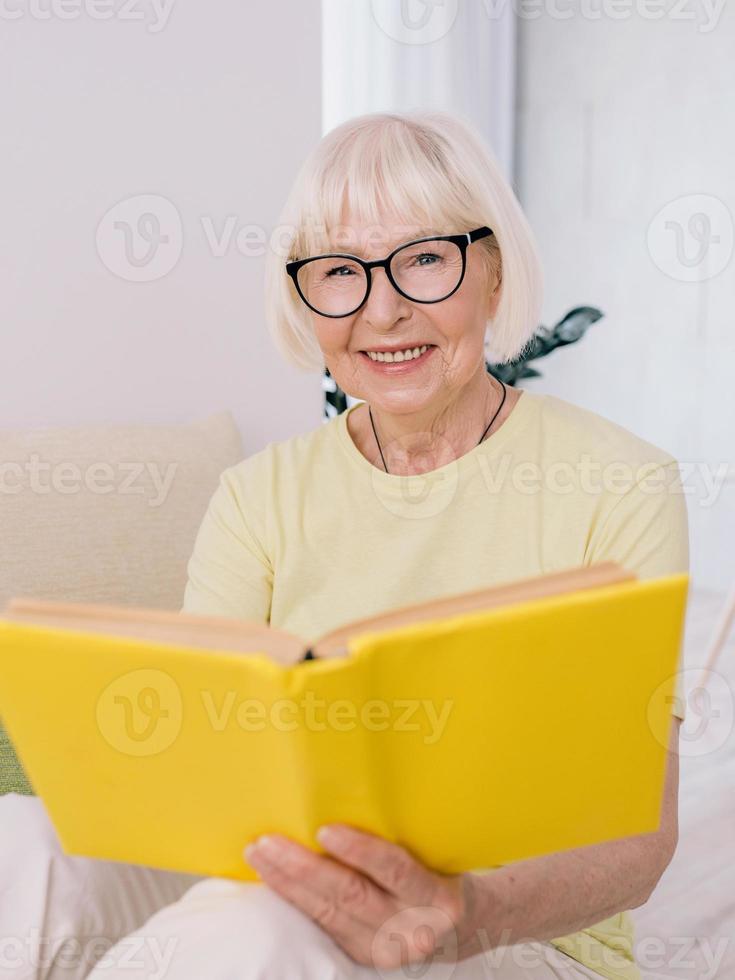 donna anziana con i capelli grigi che legge un libro su un divano a casa. educazione, pensione, anti età, concetto di lettura foto