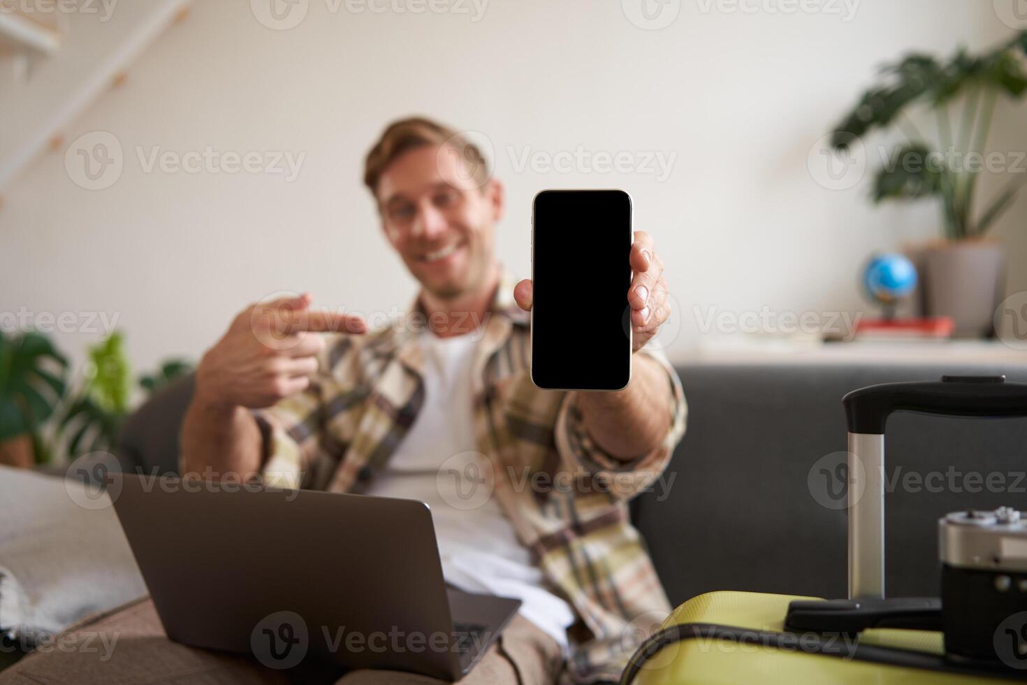 ritratto di sorridente giovane uomo, turista Spettacoli smartphone schermo, punti a mobile Telefono, raccomanda applicazione per in linea prenotazione alberghi o posto per restare su vacanza, si siede con il computer portatile foto