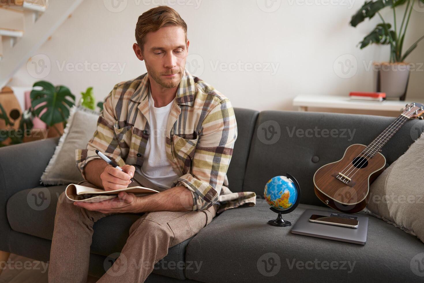 uomo scrittura lista di controllo, Imballaggio per vacanza, riferimento incrociato elementi con il suo Appunti, si siede nel vivente camera con globo e ukulele foto