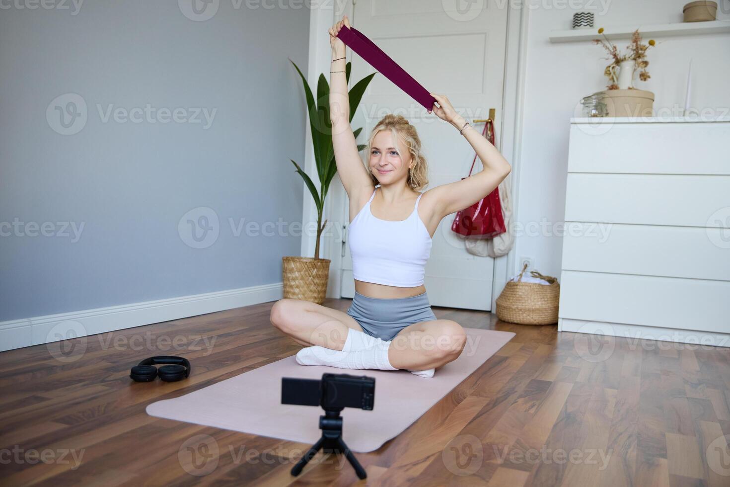 ritratto di sorridente giovane donna, la creazione di soddisfare per fitness blog, registrazione durante allenarsi con resistenza gruppo musicale, seduta su yoga stuoia foto