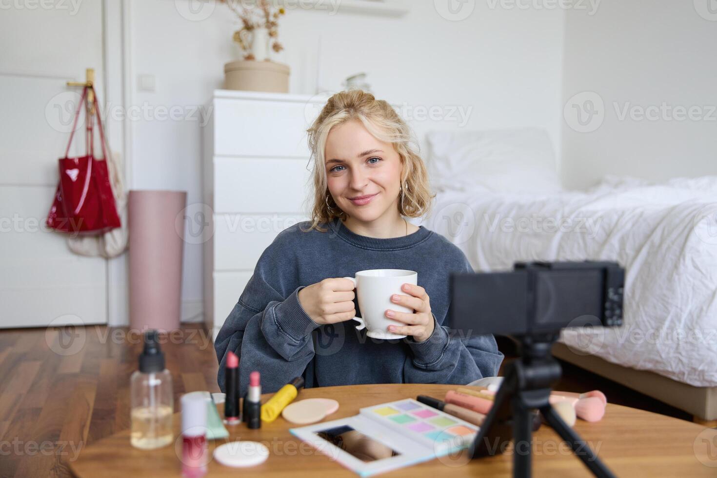 ritratto di bellissimo sociale media bellezza blogger, seduta nel davanti di digitale telecamera su pavimento nel Camera da letto, potabile tè e chattare, parlando per seguaci foto