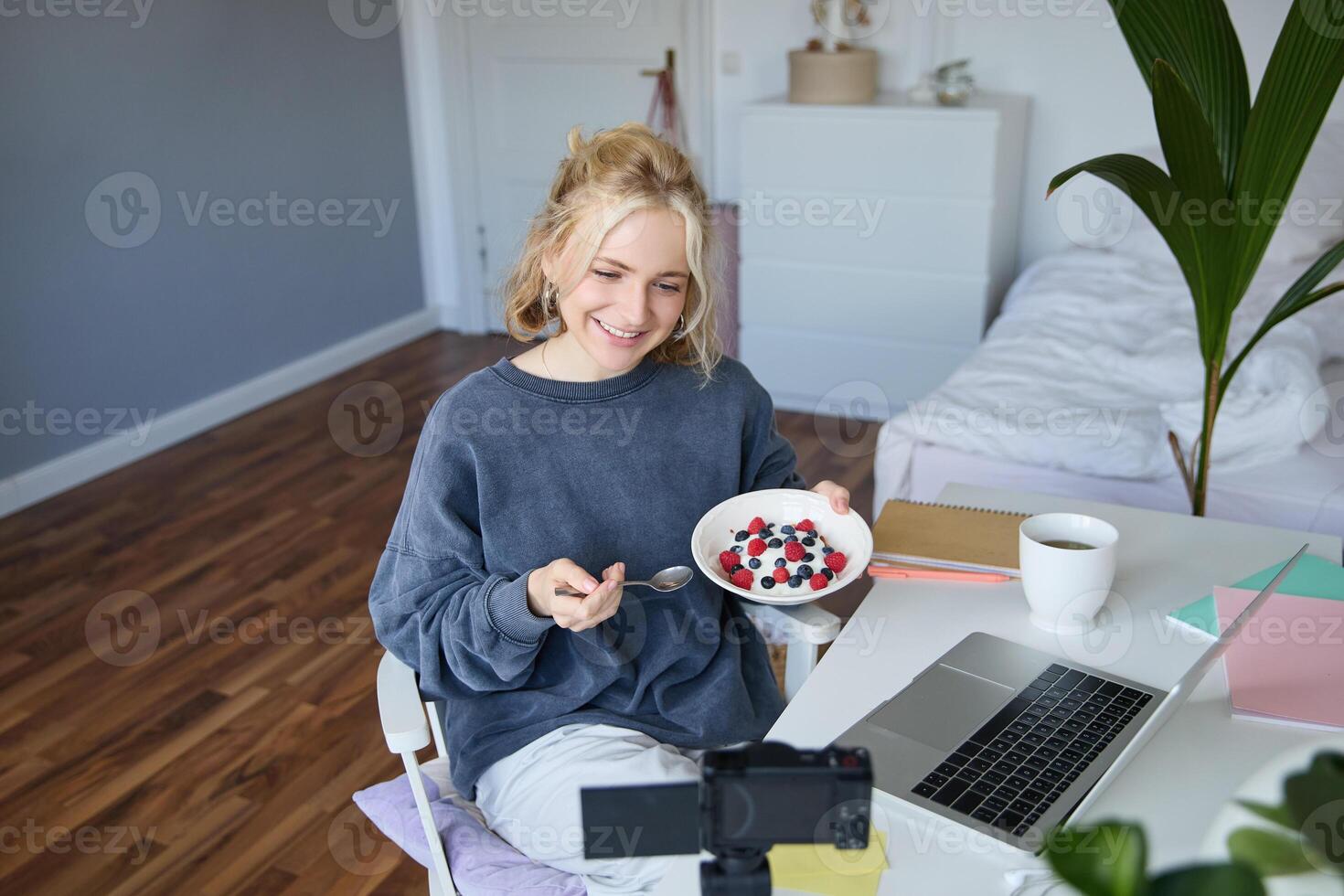 ritratto di sorridente giovane femmina vlogger, registrazione e la creazione di soddisfare nel sua camera, mangiare nel davanti di telecamera, parlando, fabbricazione stile di vita blog foto