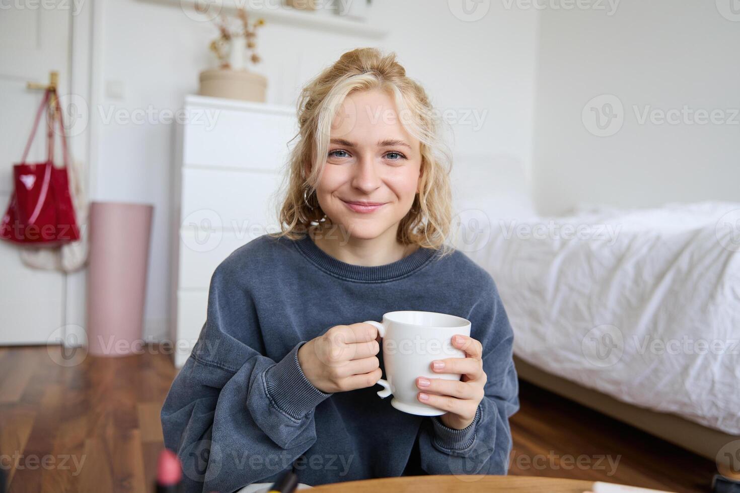 Immagine di giovane adolescenziale ragazza seduta nel sua Camera da letto su pavimento, potabile tazza di tè e godendo giorno a casa, sorridente e guardare a telecamera foto