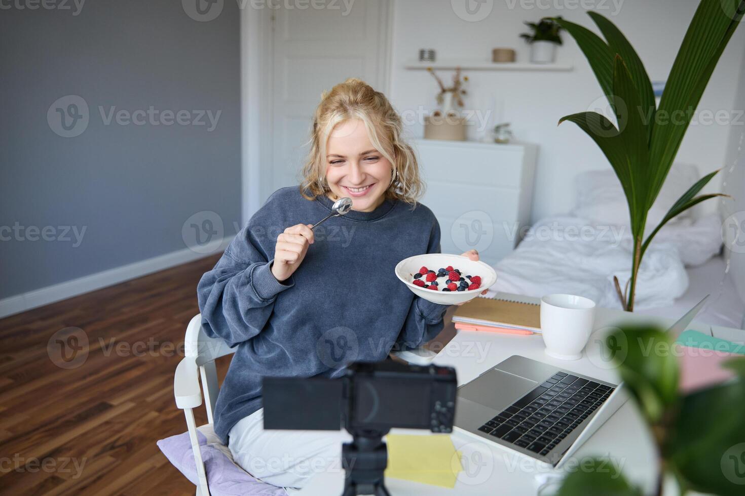 ritratto di sorridente, Candido giovane donna, soddisfare Creatore, mangiare ciotola di dolce e guardare a digitale telecamera, registrazione video blog per seguaci su sociale media foto