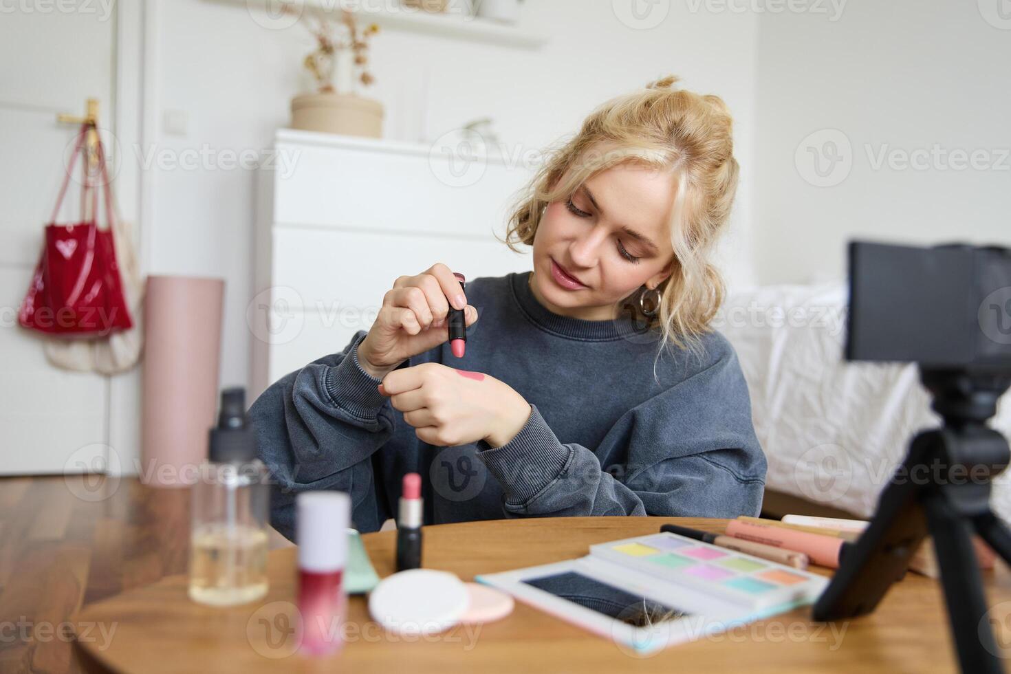 donna bellezza blogger, record di se stessa seduta nel un' camera e valutazione trucco prodotti, mette su rendere su, detiene rossetto e cosmetico spazzola nel mano, utilizzando professionale telecamera per soddisfare creazione foto