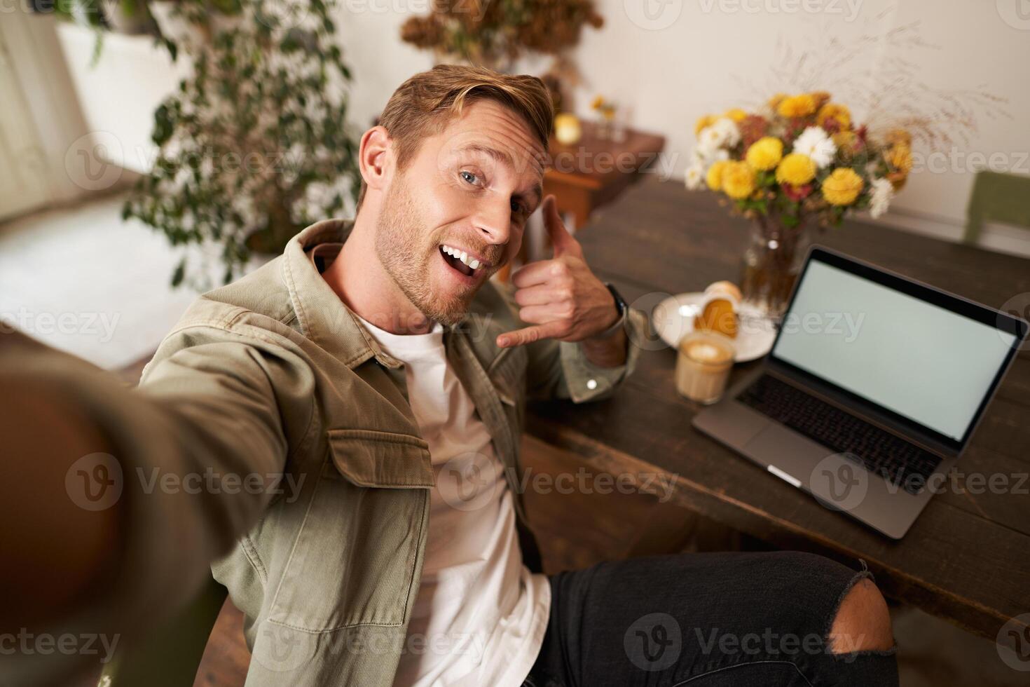 bello uomo prende autoscatto su il suo mobile Telefono con computer portatile, Spettacoli chiamata Telefono mano cartello, sorridente a telecamera, spedizione foto su sociale media app, si siede nel bar