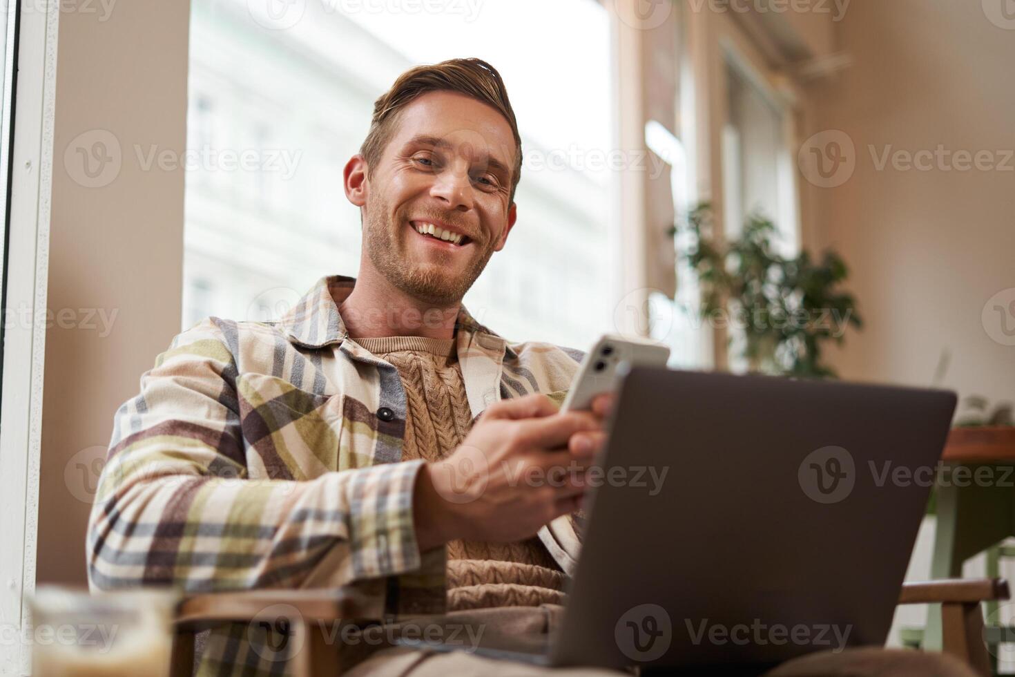 ritratto di sorridente, bello giovane uomo rilassante nel caffè negozio, Lavorando remoto a partire dal bar co-working spazio, utilizzando il computer portatile e smartphone, guardare a telecamera foto