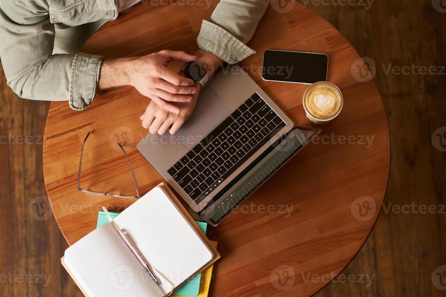 superiore Visualizza di maschio mani, uomo controllo tempo o Messaggio su il suo digitale guarda, seduta nel un' bar con computer portatile, mobile Telefono e taccuino, potabile caffè foto