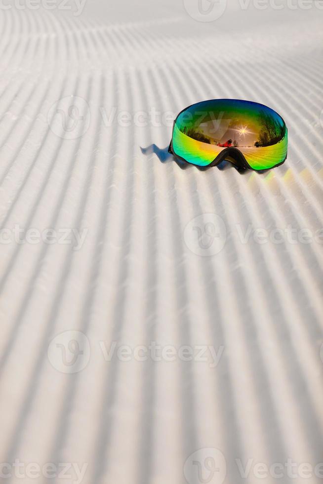 occhiali da sci posati su una nuova neve battuta e una pista da sci vuota foto