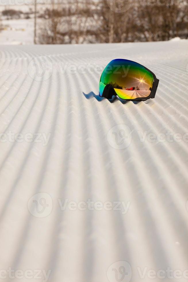 occhiali da sci posati su una nuova neve battuta e una pista da sci vuota foto