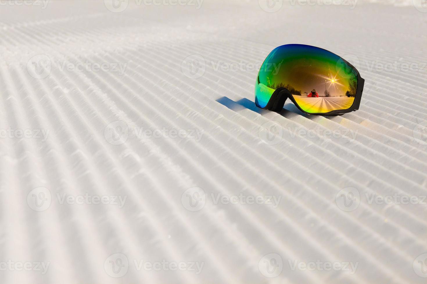occhiali da sci posati su una nuova neve battuta e una pista da sci vuota foto