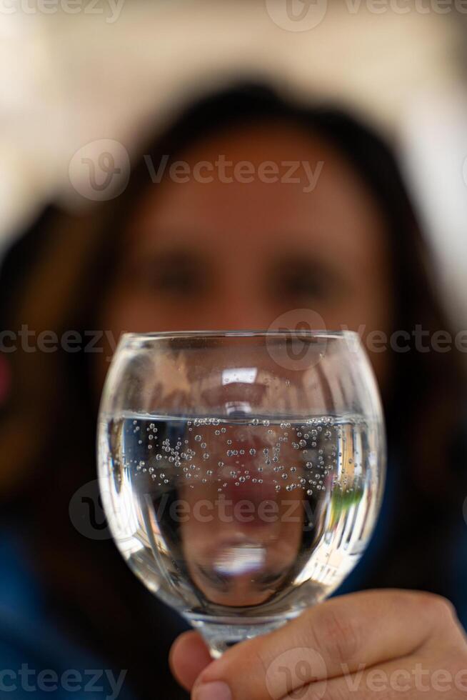 sfocato mezzo anziano donna a all'aperto ristorante mostrando sua riflessione nel un' bicchiere di acqua foto