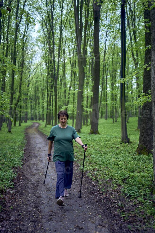 anziano donna è impegnato nel nordico a piedi con bastoni nel il primavera foresta foto