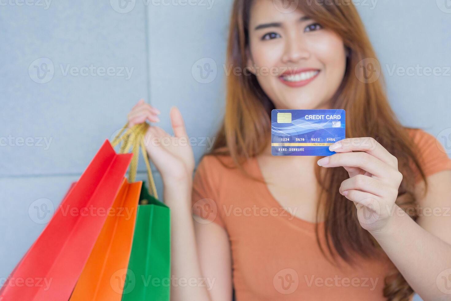 asiatico donna Tenere un' blu credito carta, colorato shopping borse, sorridente e guardare a il telecamera mentre in piedi nel il ufficio. selettivo messa a fuoco. spazio per testo. concetto di e-commerce foto