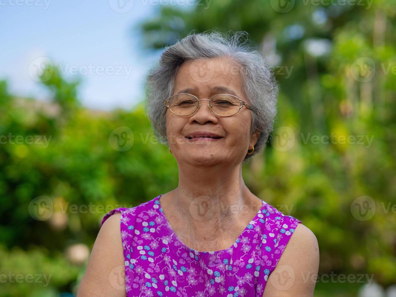 anziano donna indossare occhiali, sorridente, e guardare a il telecamera mentre in piedi nel un' giardino. spazio per testo. concetto di anziano persone e assistenza sanitaria foto