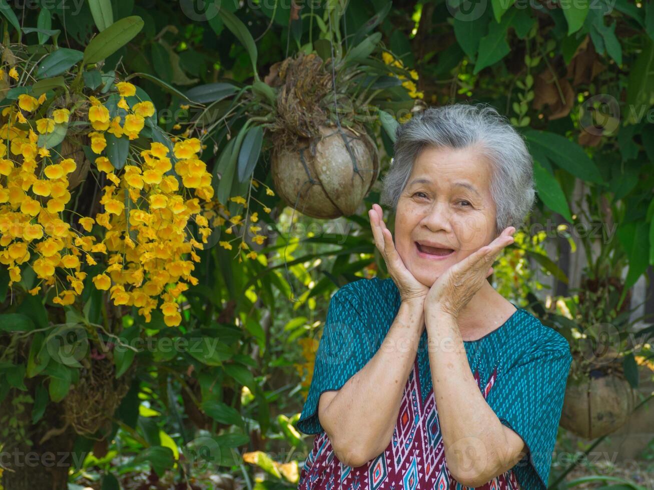 anziano donna sorridente molto contento e guardare a il telecamera mentre in piedi accanto bellissimo giallo orchidee nel un' giardino. foto