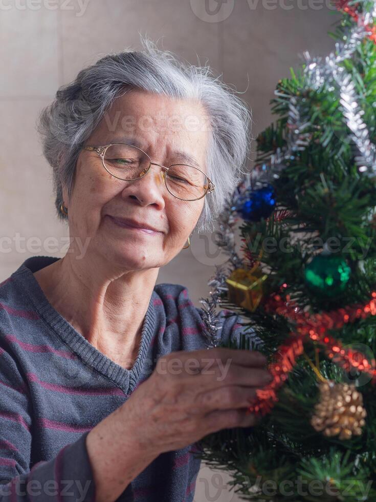 ritratto di un anziano asiatico donna decorazione il Natale albero mentre seduta nel un' vivente camera a casa. spazio per testo. concetto di anziano persone e Natale e contento nuovo anno Festival foto