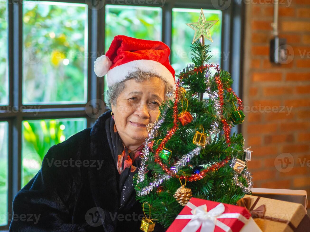 anziano asiatico donna Tenere un' regalo scatola e seduta vicino un' Natale albero a casa. concetto di anziano persone e Festival foto