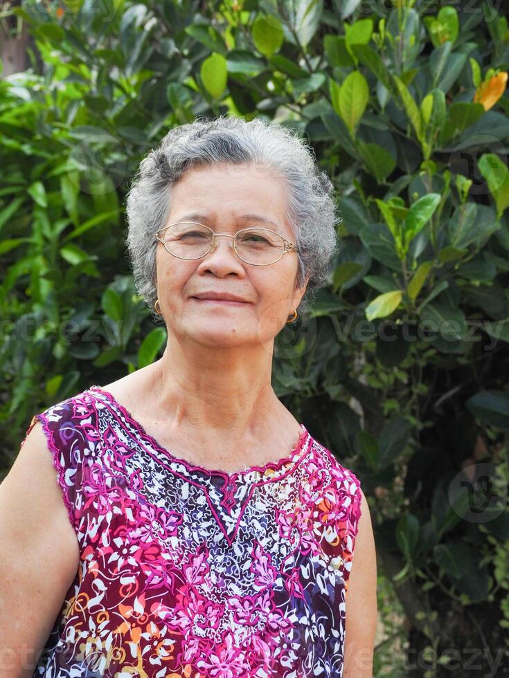 anziano asiatico donna con corto bianca capelli sorridente e guardare a il telecamera mentre in piedi nel un' giardino. spazio per testo. concetto di anziano persone e assistenza sanitaria foto