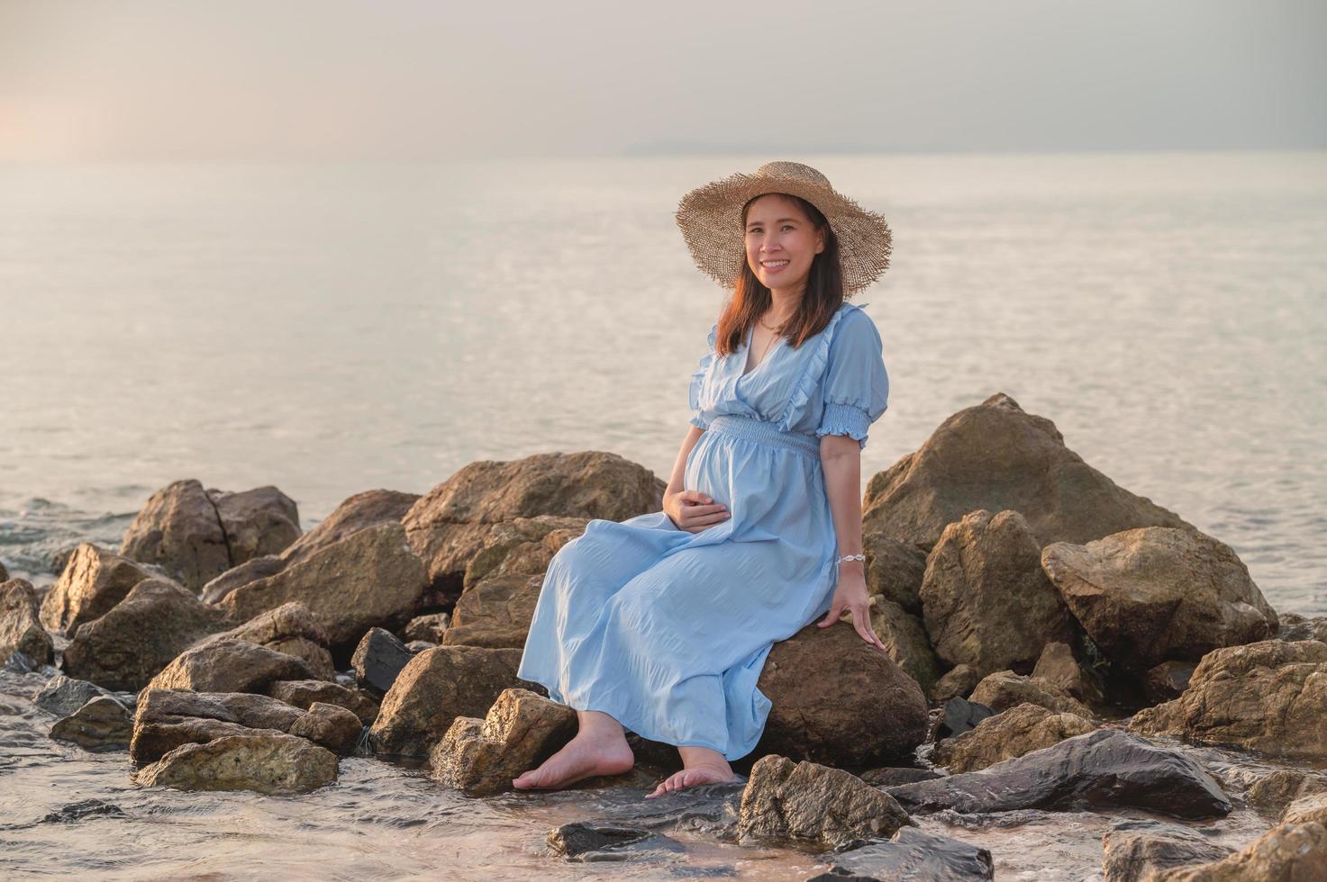 donna incinta seduta sulle rocce vicino alla spiaggia di sabbia e al mare. foto