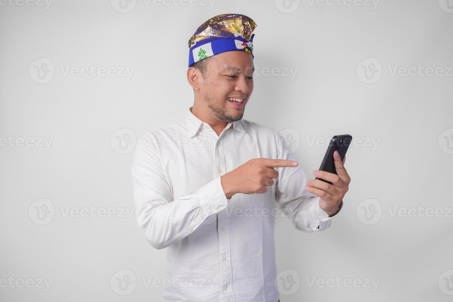 eccitato balinese uomo indossare bianca camicia e tradizionale copricapo sorridente felicemente mentre utilizzando smartphone, rispondendo Messaggio, lettura notizia foto