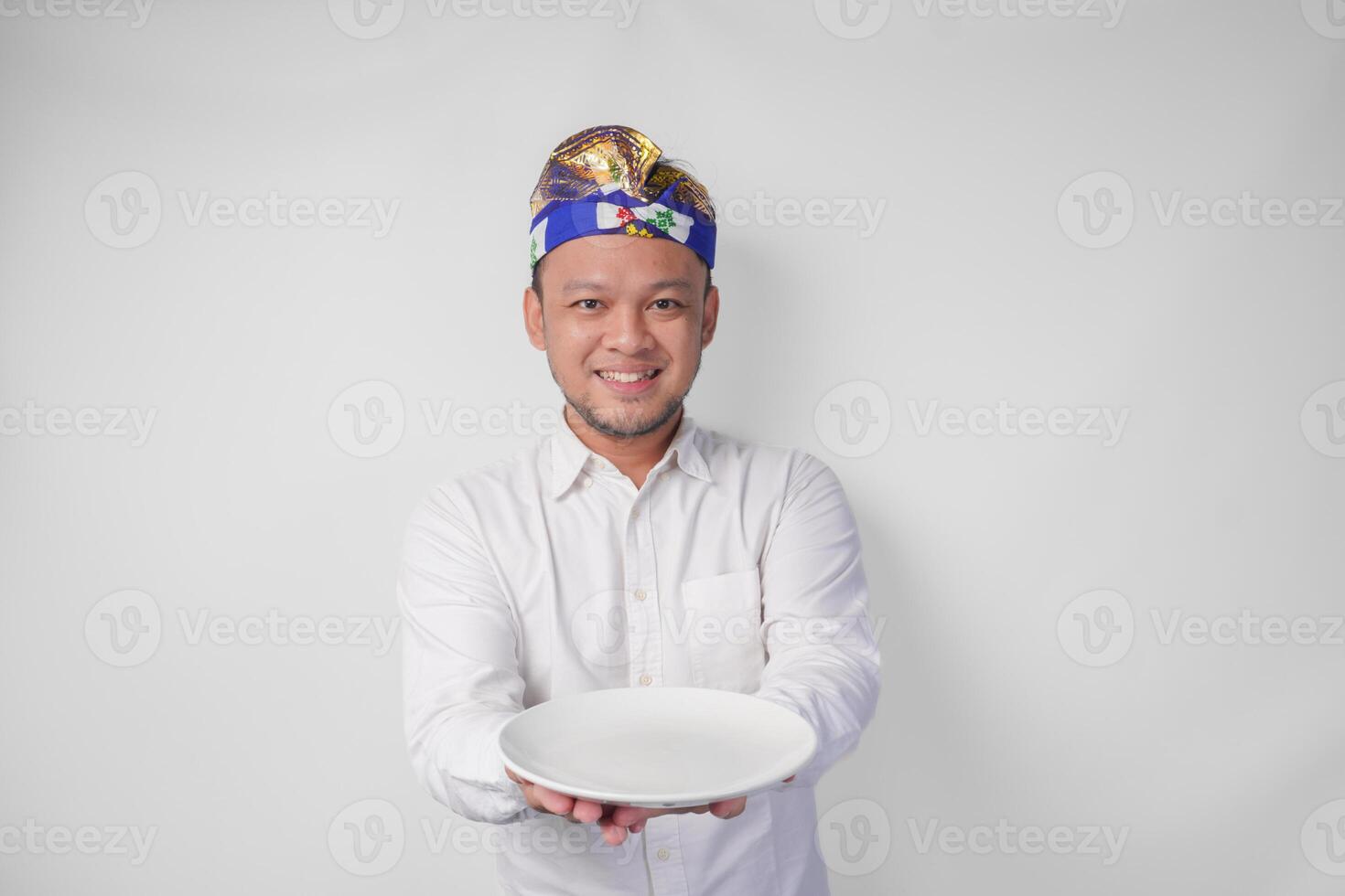 sorridente giovane balinese uomo nel bianca camicia e tradizionale copricapo mostrando e presentazione un vuoto piatto con copia spazio foto