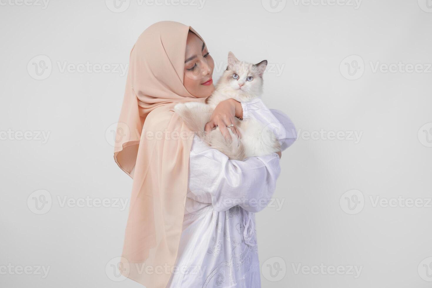 ritratto di un' bellissimo giovane musulmano donna indossare bianca vestito e hijab, sorridente e Tenere un' bianca bambola di pezza gatto al di sopra di isolato bianca sfondo foto