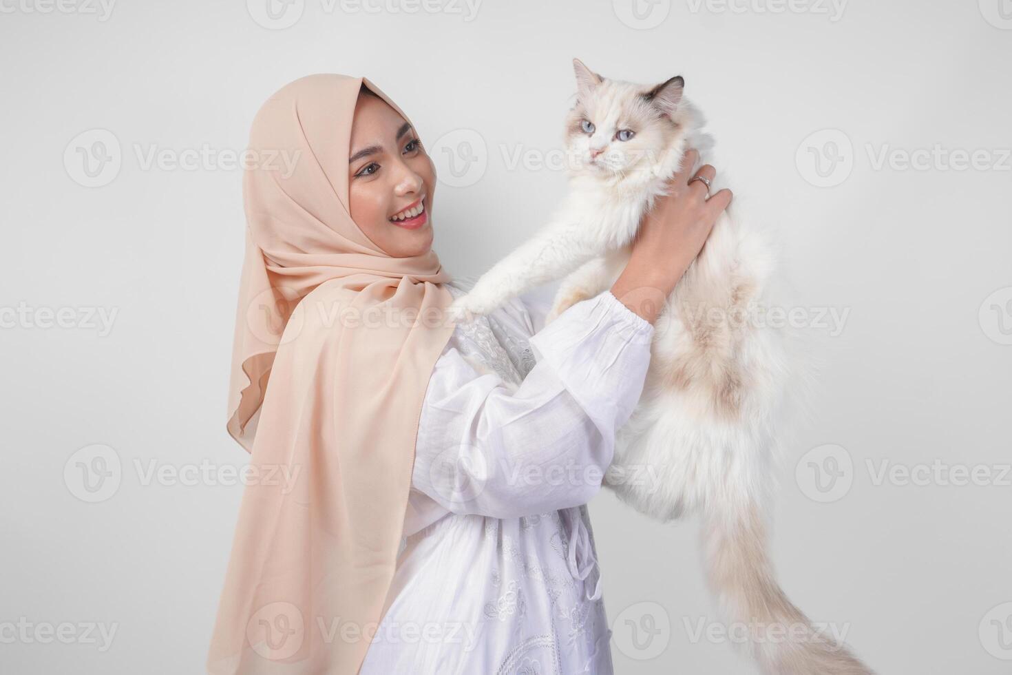 ritratto di un' bellissimo giovane musulmano donna indossare bianca vestito e hijab, sorridente e Tenere un' bianca bambola di pezza gatto al di sopra di isolato bianca sfondo foto