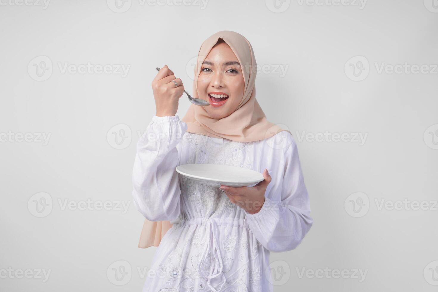 Affamato giovane asiatico musulmano donna nel hijab Tenere cucchiaio e mangiare a partire dal un vuoto piatto con copia spazio Sopra. Ramadan concetto foto