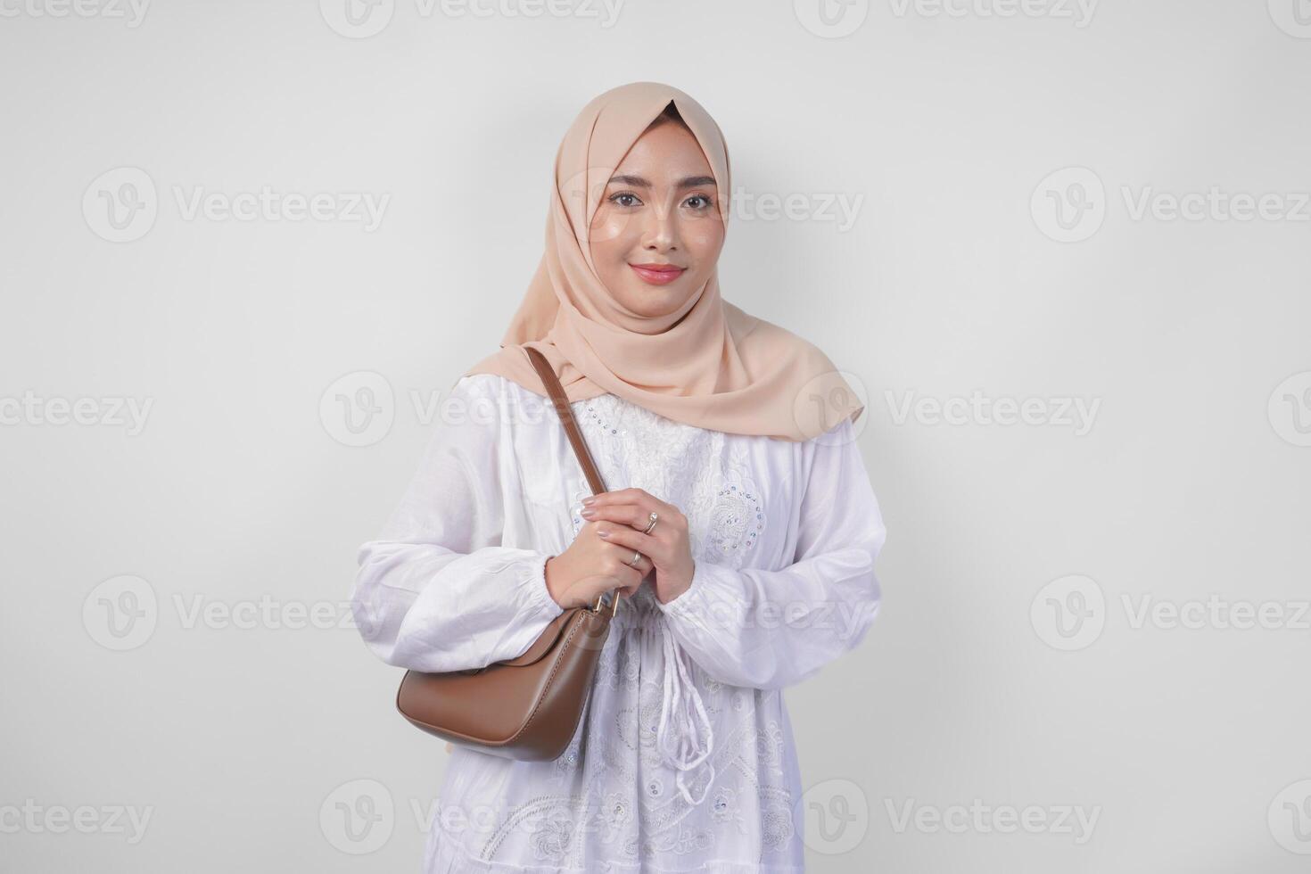 elegante giovane asiatico musulmano donna indossare bianca vestito e hijab con Marrone fionda Borsa sorridente felicemente al di sopra di isolato bianca sfondo. Ramadhan moda concetto foto