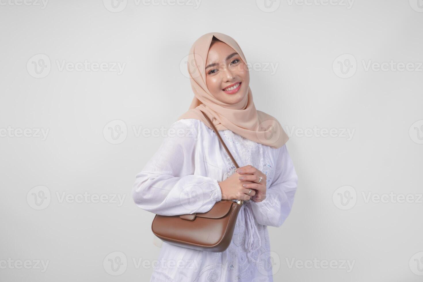 elegante giovane asiatico musulmano donna indossare bianca vestito e hijab con Marrone fionda Borsa sorridente felicemente al di sopra di isolato bianca sfondo. Ramadhan moda concetto foto