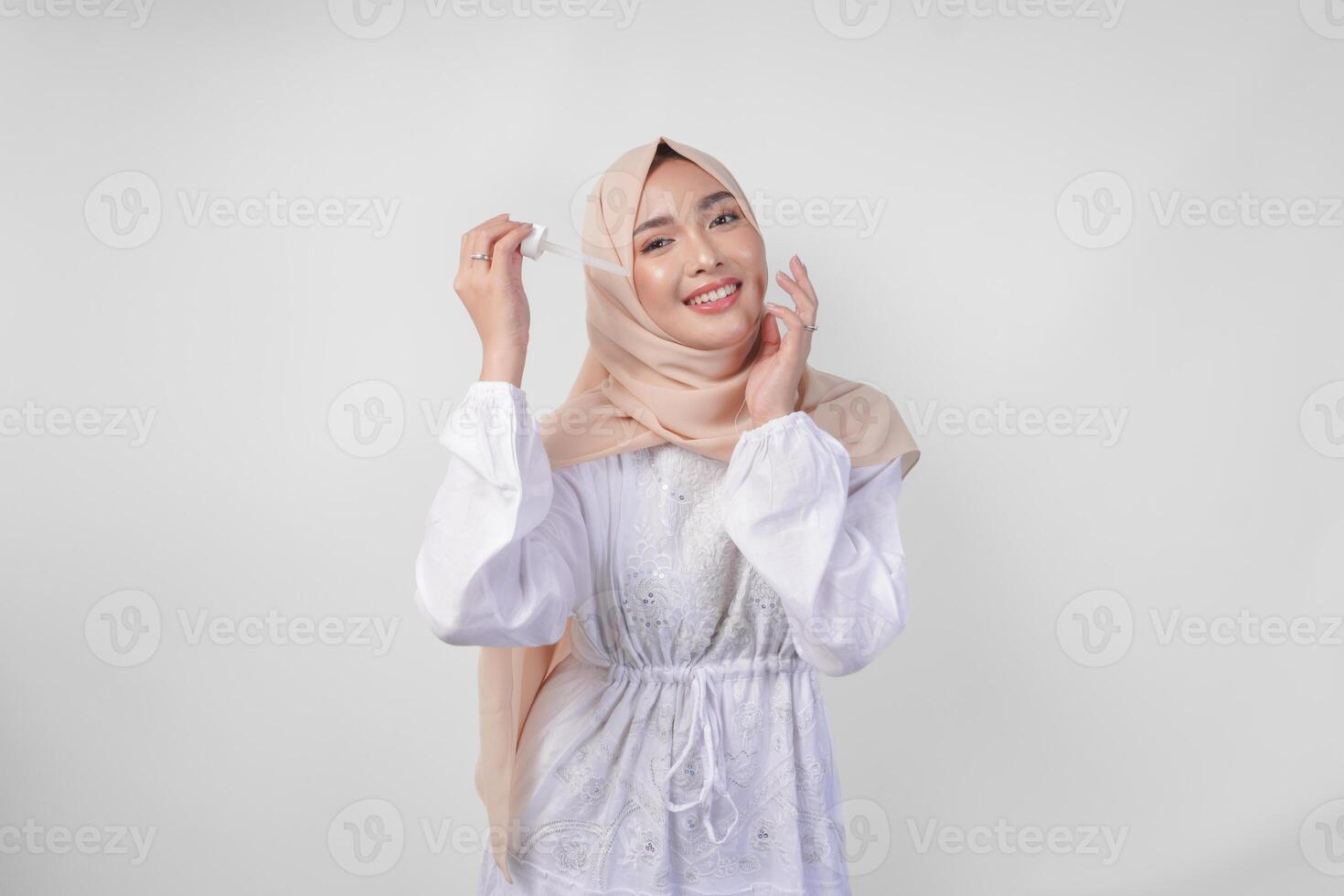 eccitato giovane asiatico musulmano donna indossare bianca vestito e hijab analisi o l'applicazione pelle cura siero su viso. facciale e bellezza concetto foto