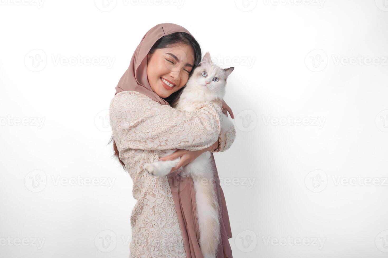 attraente giovane asiatico musulmano donna nel velo hijab sorridente mentre in posa e abbracciare un' bianca bambola di pezza gatto animale domestico foto