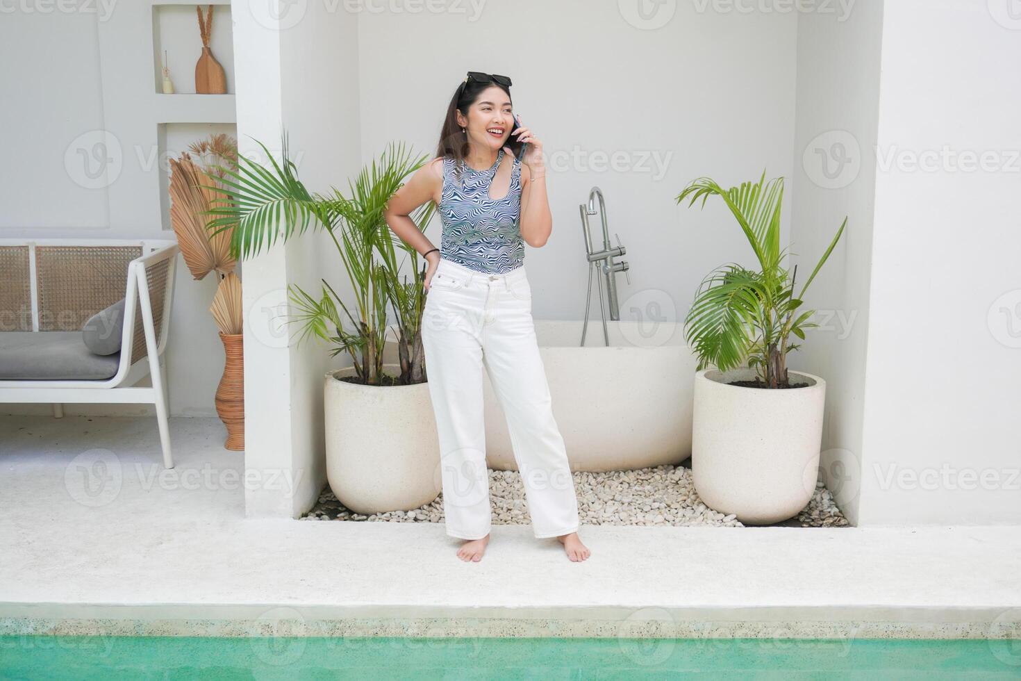 contento asiatico donna in piedi di il piscina mentre chiamata e parlando su un' mobile Telefono con un' allegro Sorridi su il viso foto