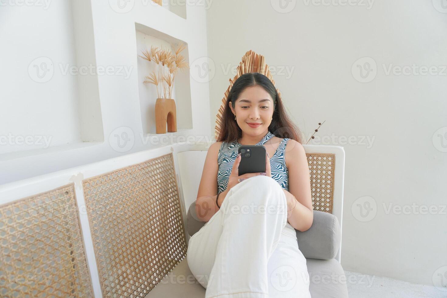 ritratto di contento asiatico donna sorridente e Tenere sua smartphone mentre posa giù su un' divano nel villa per estate vacanza foto
