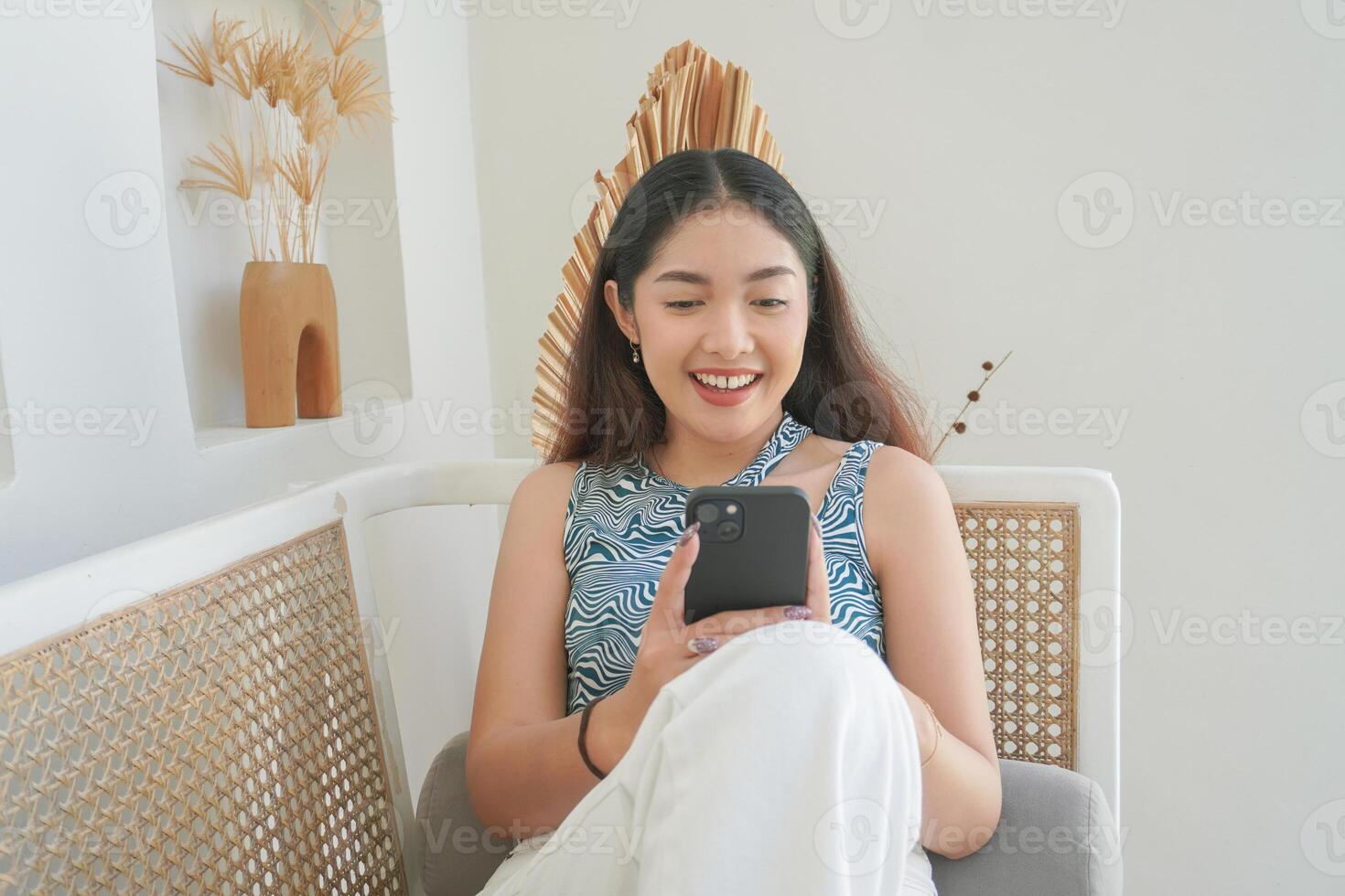 ritratto di contento asiatico donna sorridente e Tenere sua smartphone mentre posa giù su un' divano nel villa per estate vacanza foto