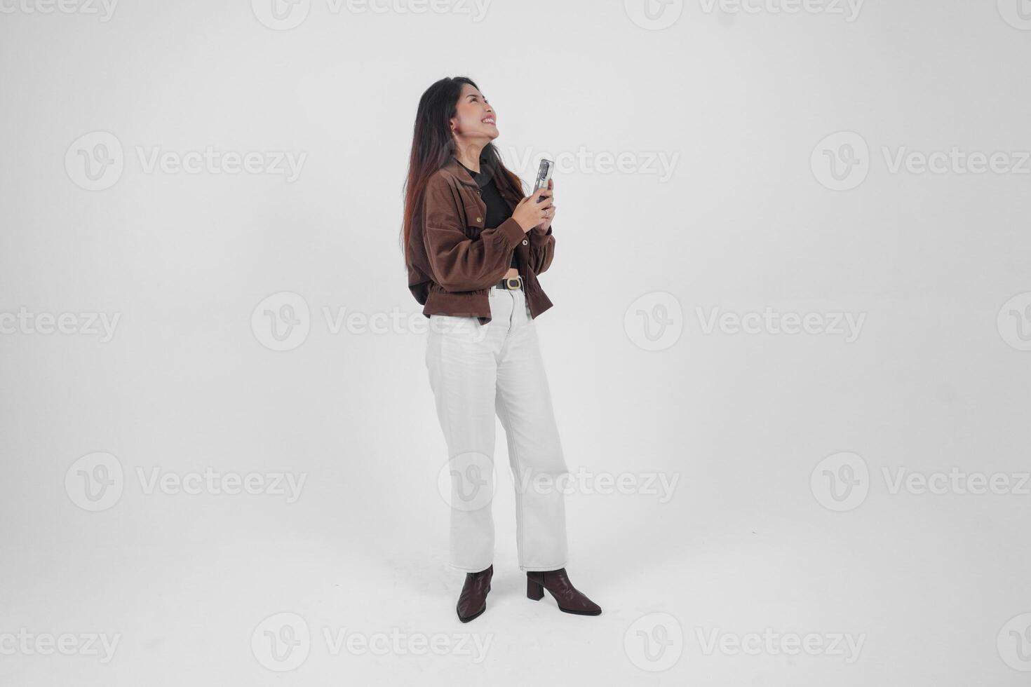 giovane asiatico donna sorridente largo guardare contento e Tenere smartphone mentre indossare casuale attrezzatura in piedi su isolato bianca sfondo foto
