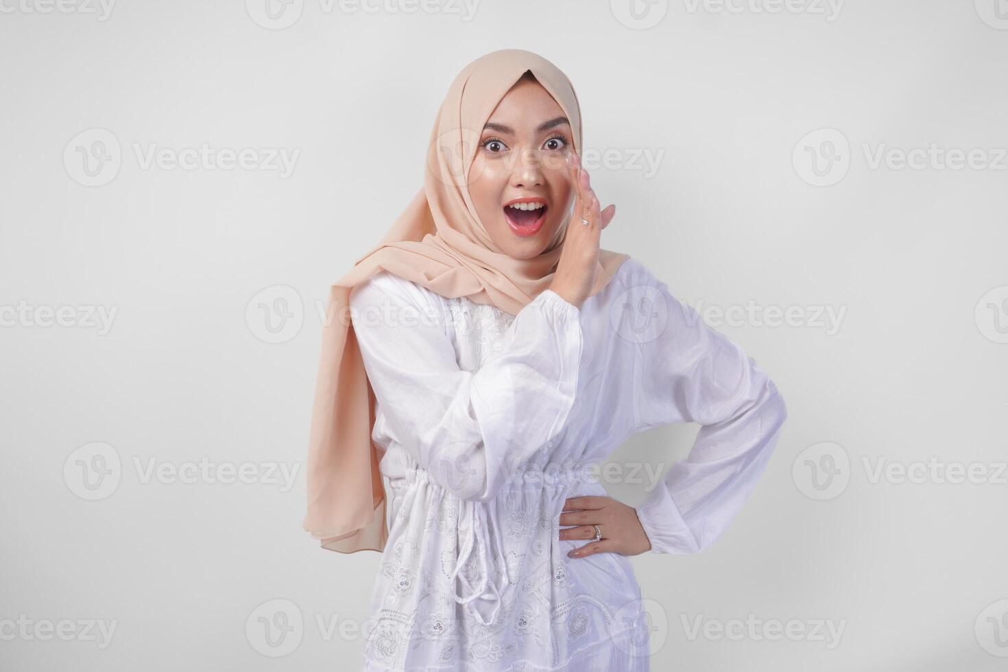 bellissimo giovane asiatico musulmano donna nel bianca vestito e hijab urlando o annunciando notizia, eccitante evento con mani su bocca, isolato su bianca sfondo foto