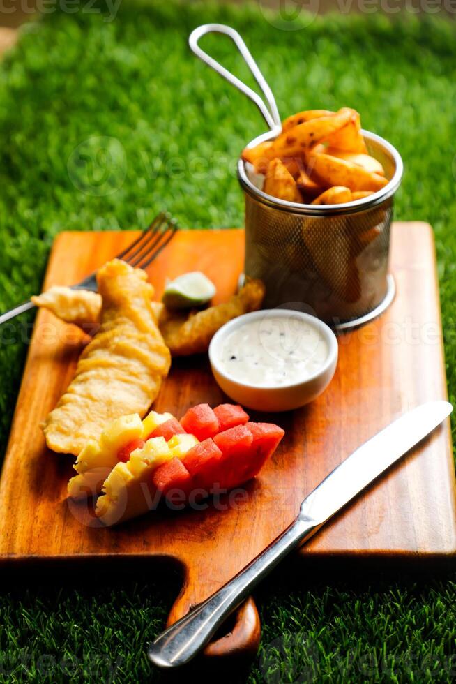 inglese pesce e patatine fritte secchio servito su di legno tavola con immersione, coltello e forchetta isolato su erboso sfondo lato Visualizza di veloce cibo foto