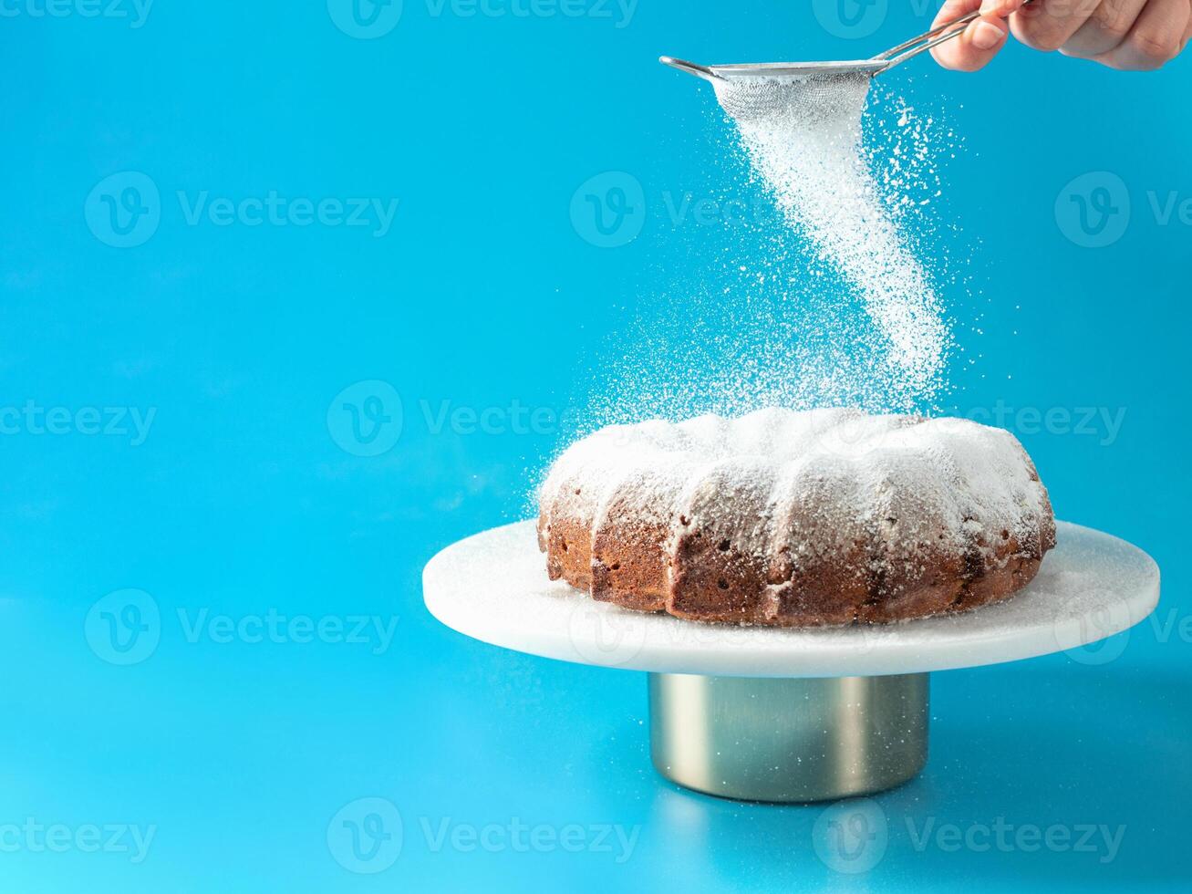 donna di mano aspersione glassatura zucchero al di sopra di fresco casa fatto bundt torta. polvere zucchero cascate su fresco Perfetto bunt torta al di sopra di blu sfondo. copia spazio per testo. idee e ricette per prima colazione o dolce foto